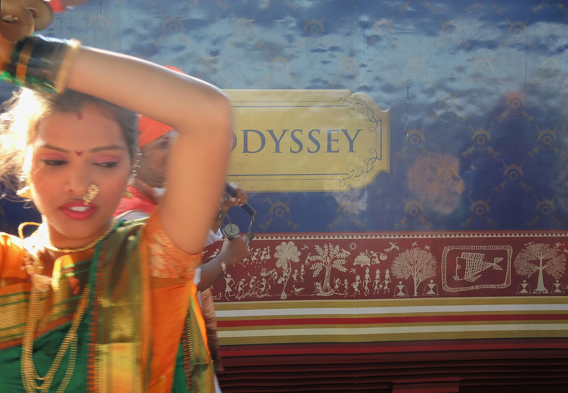 A folk dancer welcoming guests beside the blue, Warli-painted livery of the Deccan Odyssey