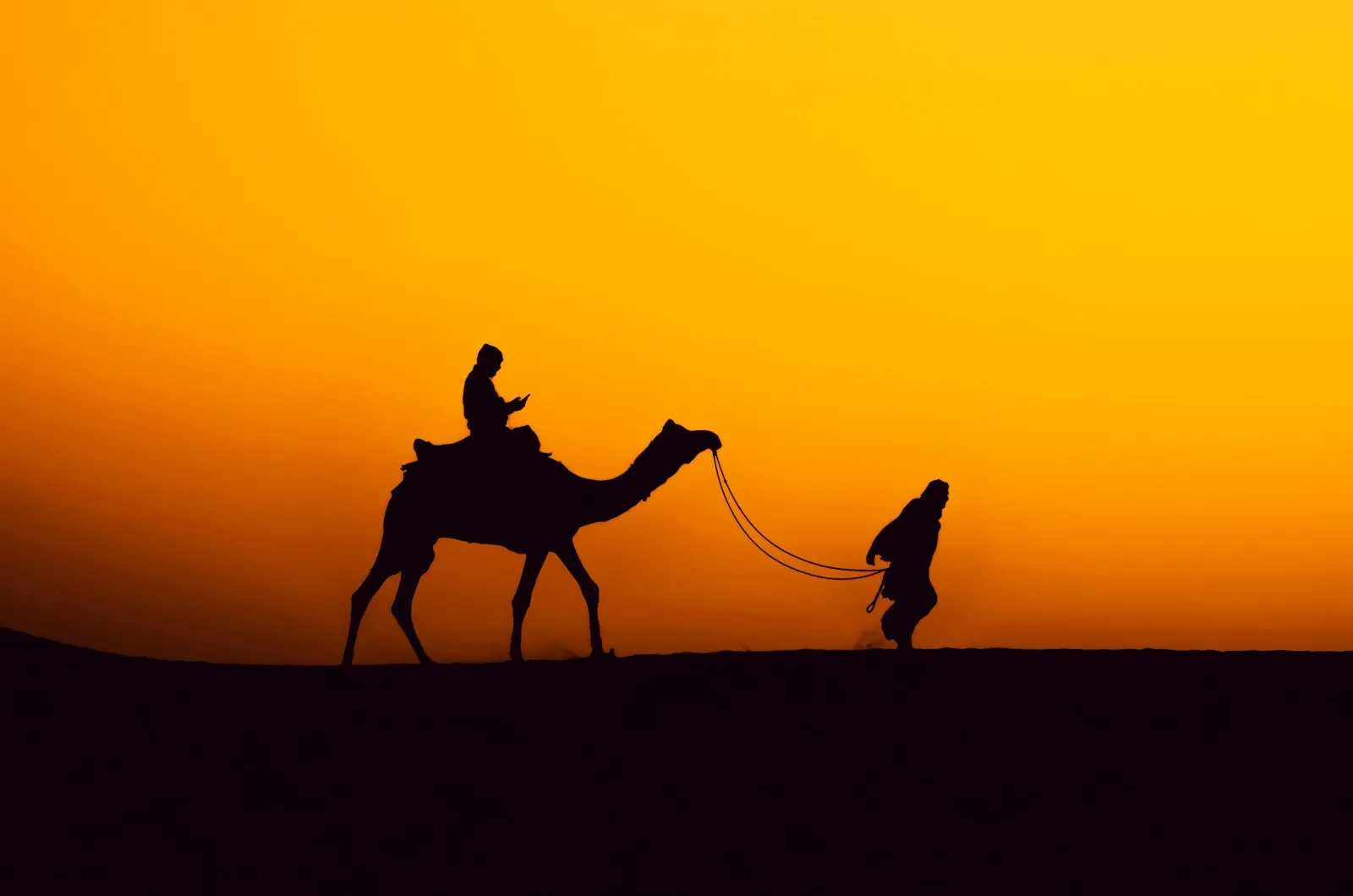 A camel and riders silhouetted against a blazing desert sunset near Jaisalmer