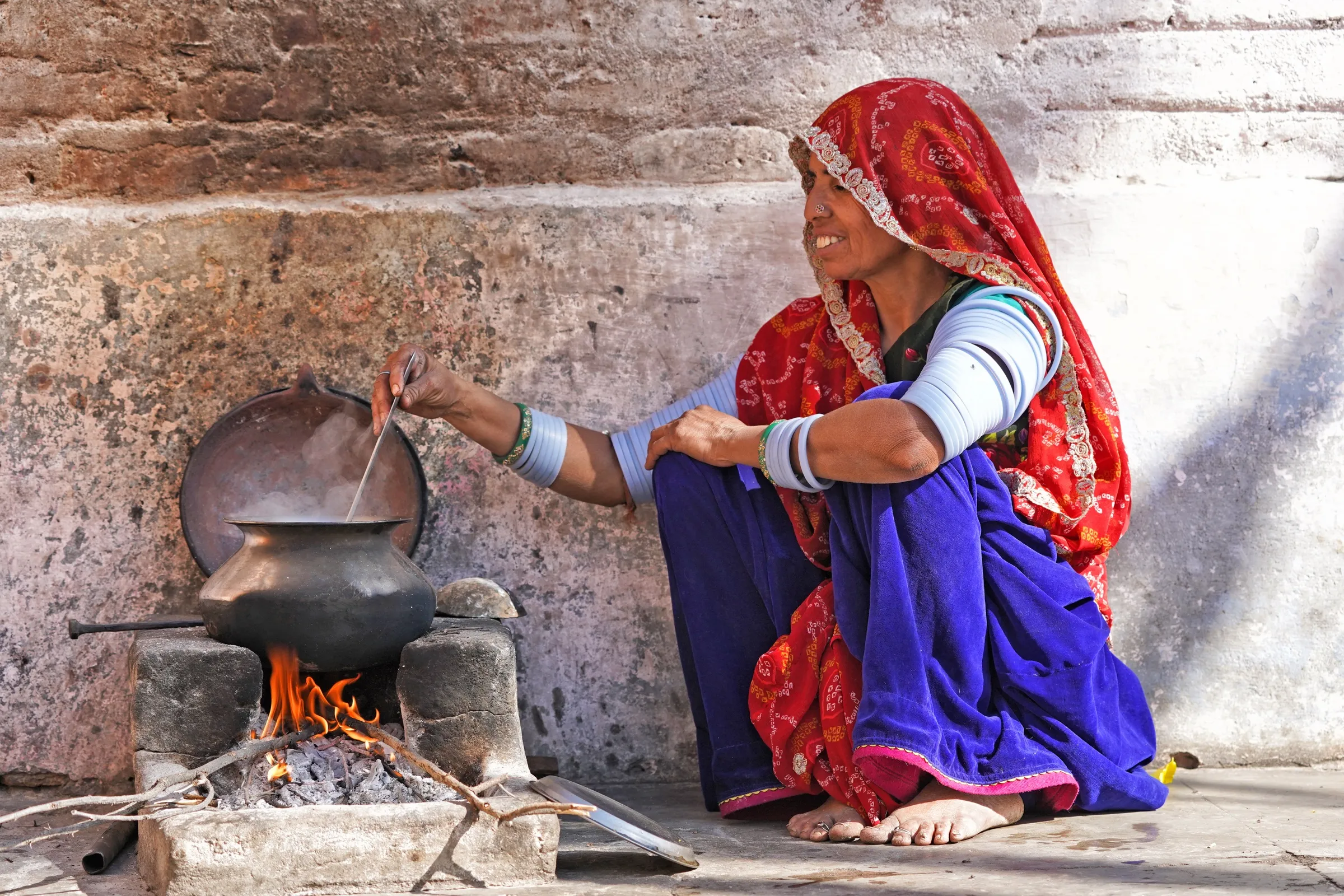 Woman in a red veil cooking over an open fire beside a stone wall