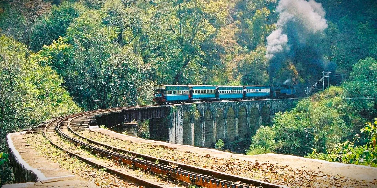 A Nilgiri Mountain Railway train crossing a stone viaduct through green hills towards a tunnel