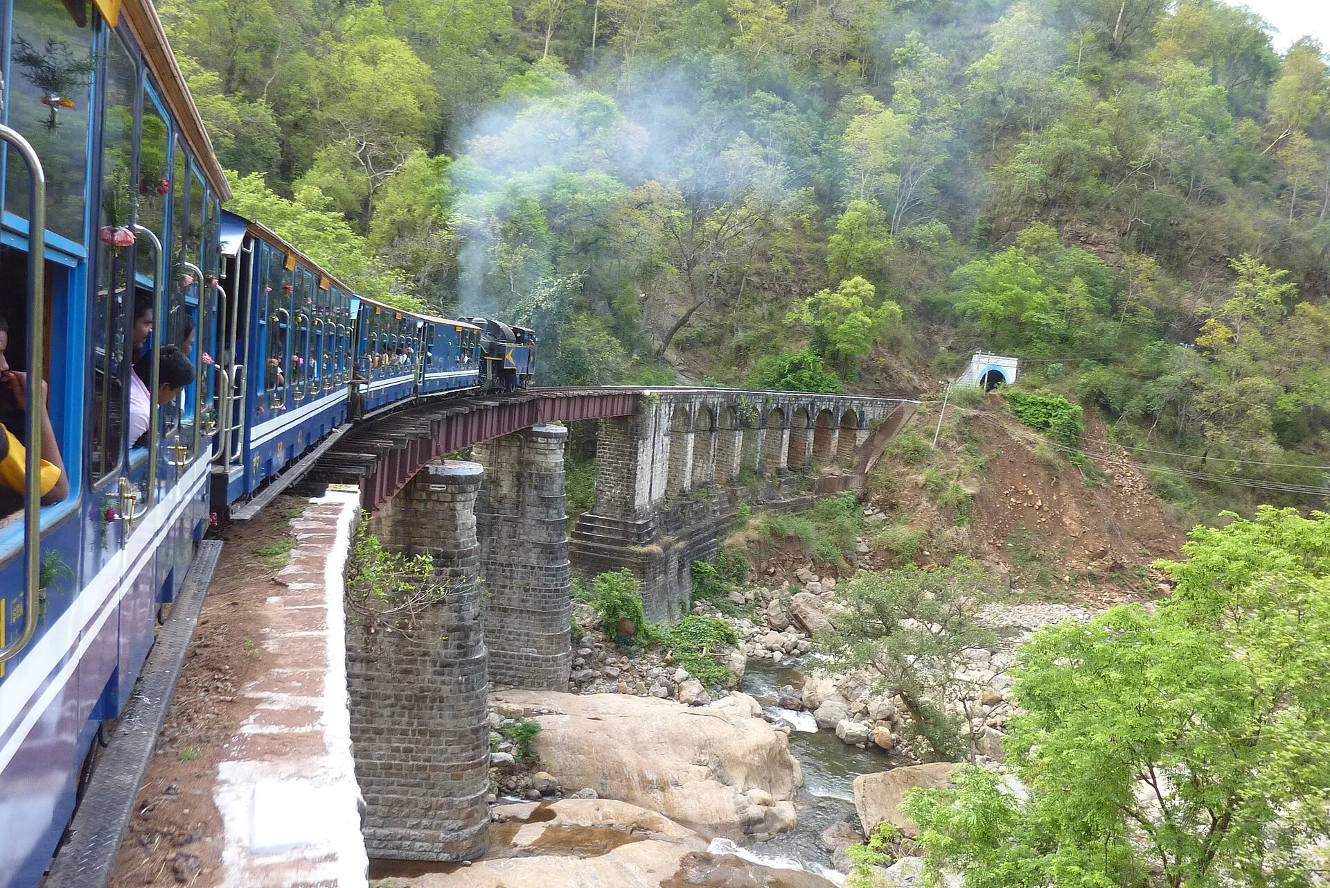 A Nilgiri Mountain Railway train crossing a stone viaduct through green hills towards a tunnel